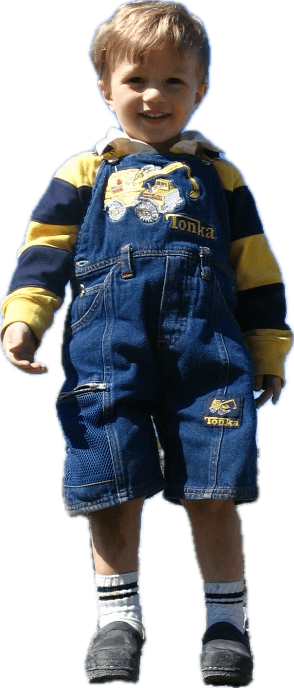 A full shot cutout of a smiling young boy with blonde hair. He wears denim short overalls with a Tonka truck graphic and logo, a striped yellow and dark blue shirt, white socks with black stripes, and dark clogs.